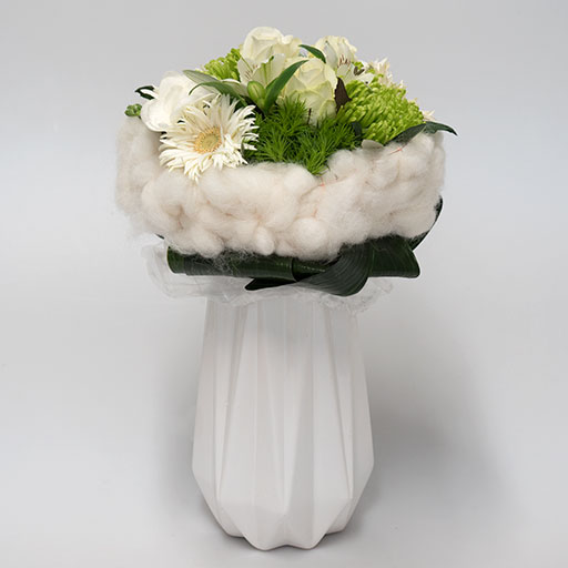 Ein Neststrauß mit weißen und grünen Blumen, umringt von einem Nest aus weißer Watte, in einer weißen Vase.